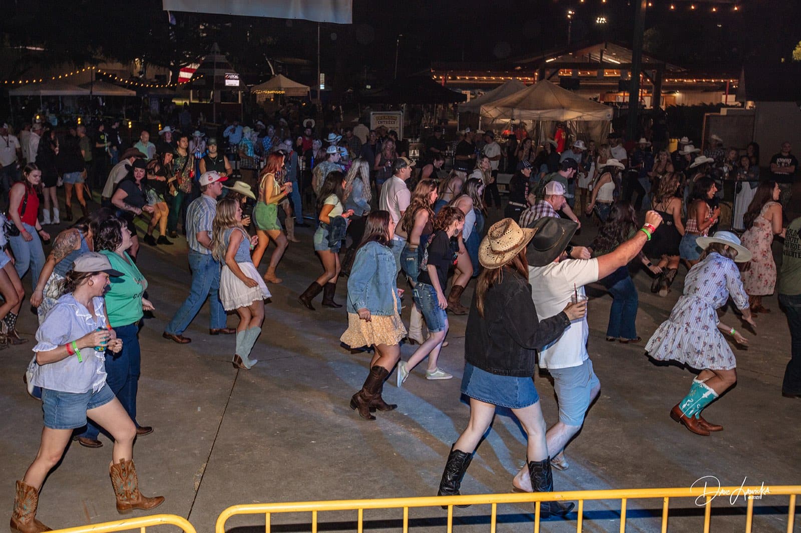 Crowd in cowboy boots and hats line dancing under string lights at the Barn Dance Bash after party