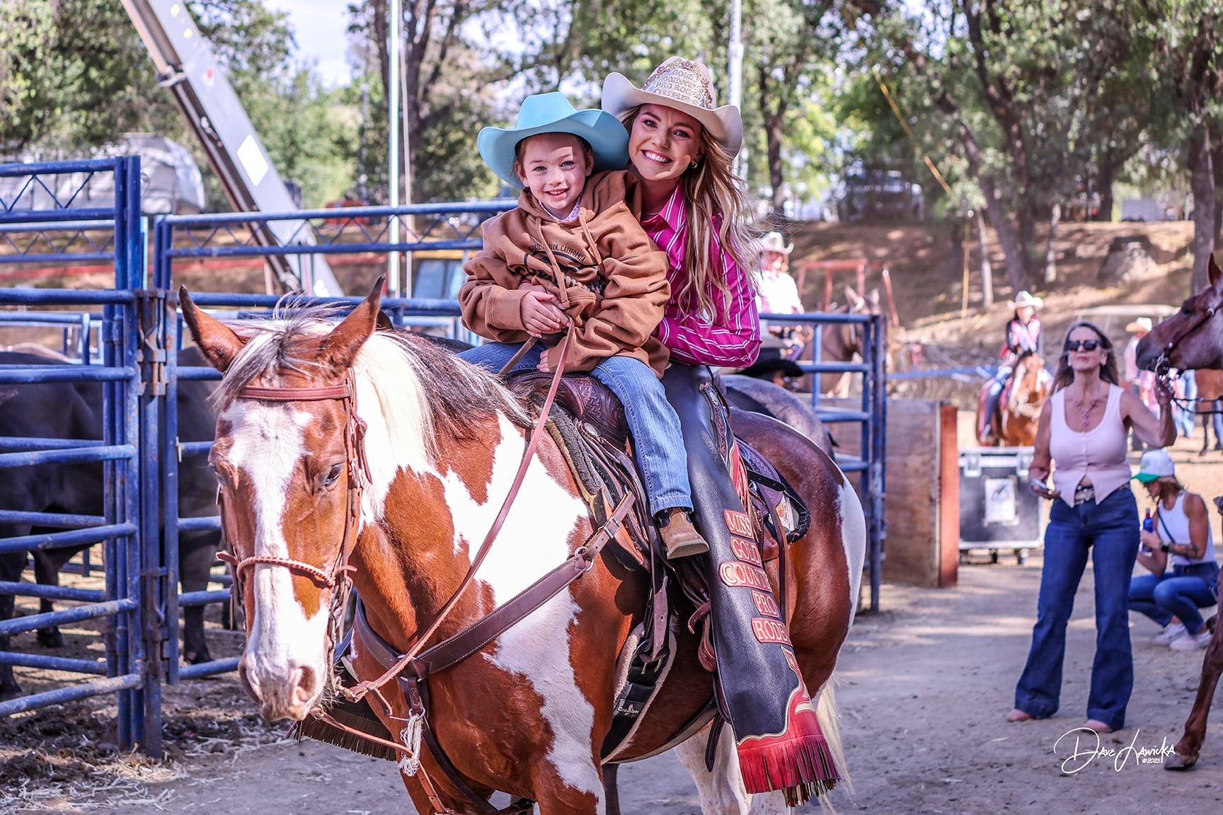 Smiling child in a turquoise cowboy hat riding a paint horse with a cowgirl in a pink shirt at the Gold Country Pro Rodeo