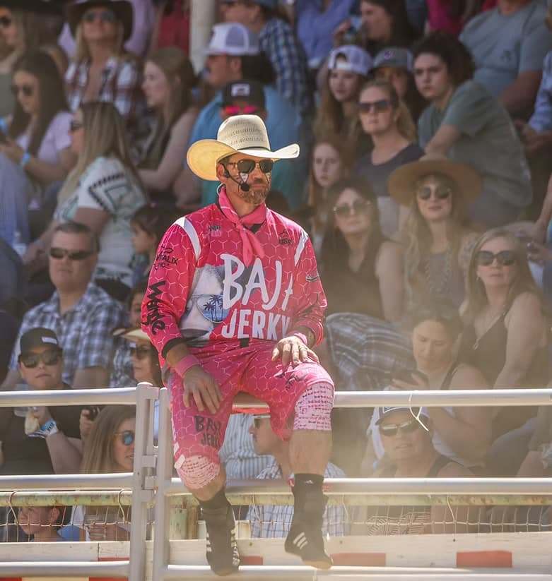 Rodeo entertainer in a pink suit and cowboy hat sitting on the arena rail in front of the crowd